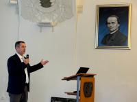 A man in a jacket speaking into a microphone gestures. A framed picture of Gregor Mendel hangs on the wall.
